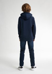 Mörkblå hoodie med känguruficka och ribbade ärmslut, kombinerad med mörkblå jeans och vita sneakers med svarta detaljer.
