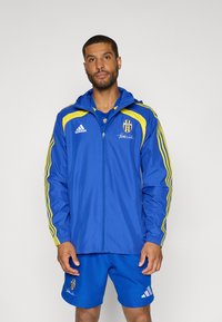 Blauwe windjack met gele accenten, ritssluiting en capuchon. Heeft drie strepen op de mouwen en een geborduurd logo. Gecombineerd met blauwe korte broek.