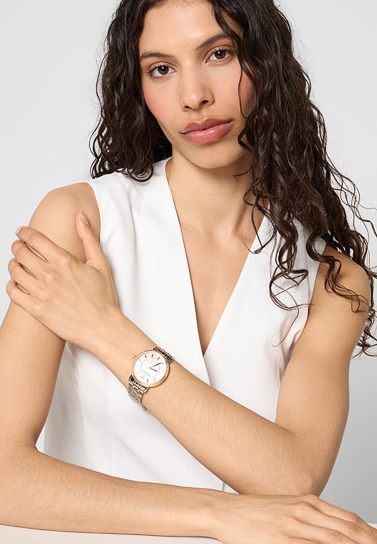 Montre dorée et argentée avec un cadran rond blanc, bracelet en maille et accents en cristal autour de la lunette. Portée sur un poignet nu.