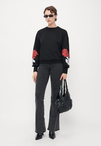 Person med svart hettegenser med rød og hvit blomsterdesign på ermene, mørke grå jeans, svarte støvler, solbriller, og holder en svart håndveske.