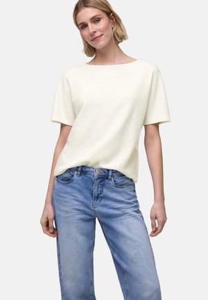 Junge Frau mit kurzen blonden Haaren, die ein cremefarbenes, geripptes Kurzarmtop und hellblaue, hoch taillierte Jeans trägt und mit einem leichten Lächeln dasteht.