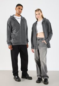 Felpa grigia con zip e cappuccio, pantaloni baggy neri e grigi e scarpe con platform nere. Fit casual, texture a contrasto e dettagli di cucitura sottili.