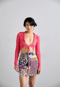 Červený rebrovaný crop top s dlhými rukávmi a viazaním vpredu, v kombinácii s miniskirtou z patchworku v rôznych farbách, ktorá má kvetinové a geometrické vzory.