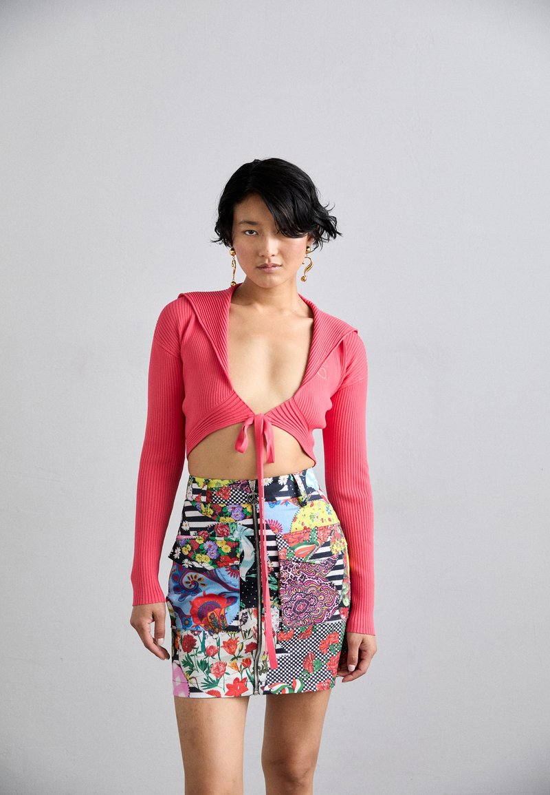 Červený rebrovaný crop top s dlhými rukávmi a viazaním vpredu, v kombinácii s miniskirtou z patchworku v rôznych farbách, ktorá má kvetinové a geometrické vzory.