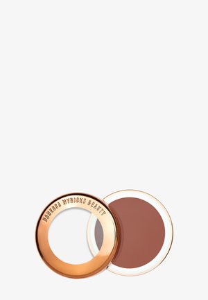 Boîte de maquillage ronde avec un couvercle rose doré scintillant embossé de "Danessa Myricks Beauty" et un produit crème marron à l'intérieur.