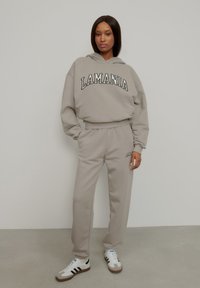 Šedá mikina so potlačou "LAMANIA", doplnená o zodpovedajúce teplákové nohavice. Tkanina vyzerá jemne a štruktúrovane. Bielozelené a čierne tenisky dotvárajú celkový vzhľad.