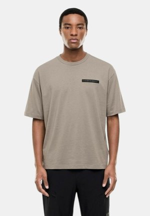Giovane uomo con capelli corti neri che indossa una t-shirt oversize color taupe con piccolo logo nero sul petto e pantaloni neri, in piedi davanti a uno sfondo semplice.