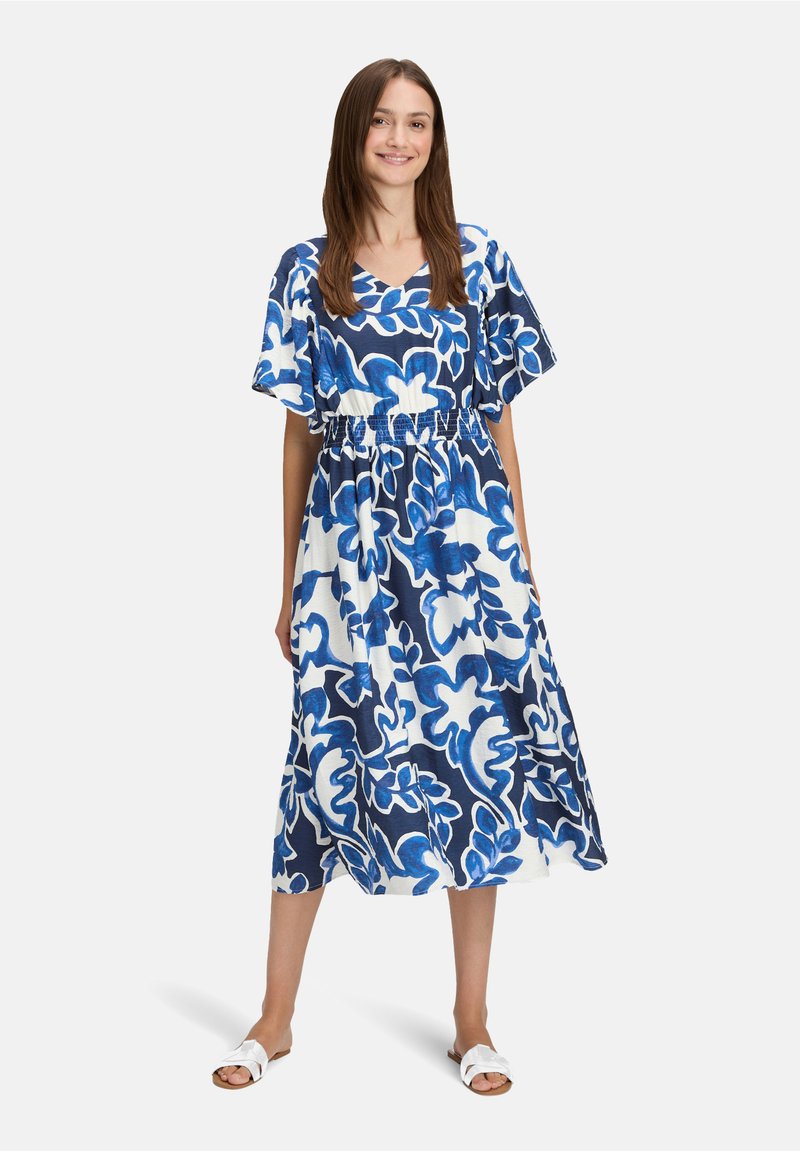 Femme debout souriante, portant une robe midi à motifs floraux bleus et blancs avec manches courtes et sandales plates blanches.