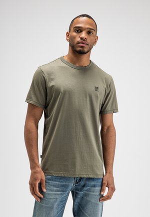 Mann steht vor schlichtem Hintergrund, trägt ein olivgrünes T-Shirt mit kurzen Ärmeln und blaue Jeans, schaut leicht zur Seite.