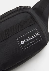 Sort stof bumbag med en tekstureret overflade, der featuring en lynlås, justerbar rem og et hvidt logo-applikation med "Columbia" tekst.