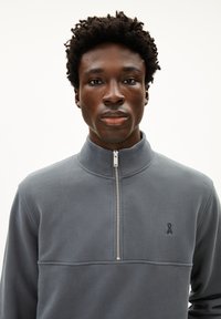 Felpa zip-up grigio scuro in pile con colletto alto, caratterizzata da un piccolo logo ricamato sul petto e dettagli di cucitura visibili.