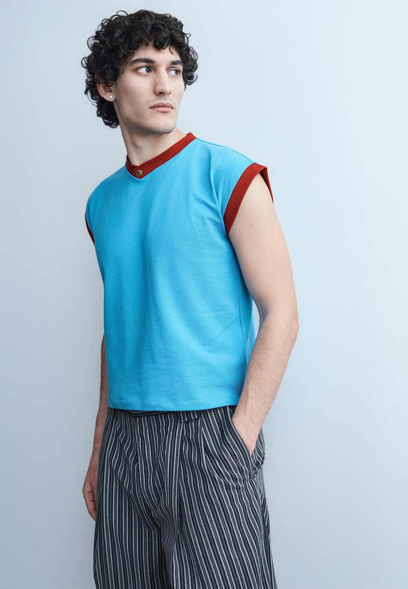 Young man with curly dark hair wearing a sleeveless blue shirt with red trim and striped black and white pants, looking to the side.