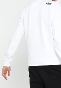 Person i vit långärmad sweatshirt med "The North Face"-logotyp på den övre ryggen, i sällskap med svarta byxor, vänd bort.