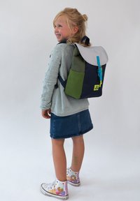 Batoh v námorníckej modrej, zelenej a svetlosivej farbe, so modrým zipsom, zapínaním na klopu a postrannou kapsou. Dieťa v svetlom svetri a džínsovej sukni.