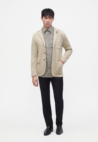 Jeune homme portant un blazer beige par-dessus une chemise à carreaux, un pantalon noir et des chaussures noires, debout avec une main dans la poche sur fond blanc.