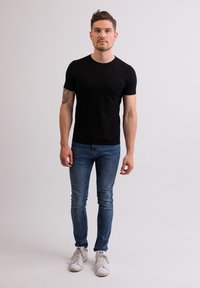 Černé tričko s krátkým rukávem, těsně přiléhavého střihu, spojené s modrými slim-fit džíny a bílými teniskami, stojící na jednobarevném pozadí.
