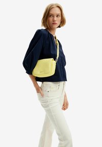 Borsa a tracolla gialla con motivo a stella testurizzato; presenta una catena e una tracolla regolabile. Indossata con una blusa blu navy e jeans bianchi.