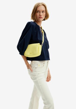 Gelbe Umhängetasche mit texturiertem Sonnenstrahlenmuster; verfügt über eine Kette und einen verstellbaren Riemen. Getragen mit einer marineblauen Bluse und weißen Jeans.