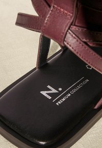 Bordeaux lædersandal med en flad sort sål. Har krydsede stropper og "N. PREMIUM COLLECTION" logo trykt på indersålen.