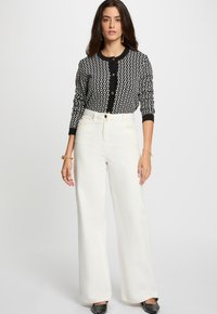 Gilet à motifs noir et blanc avec un col rond, fermeture par boutons et manches longues ; associé à un pantalon blanc à taille haute et jambes larges.