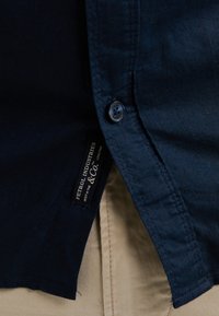 Chemise bleu marine à manches courtes avec col boutonné, tissu texturé et étiquette de marque visible sur la couture latérale.