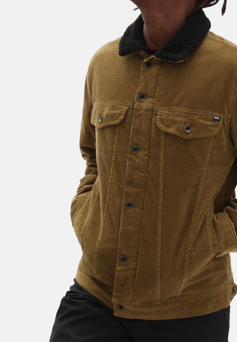 Personne portant une veste en velours côtelé marron avec un col en polaire noire et un pantalon noir, mains dans les poches de la veste, sur un fond blanc uni.