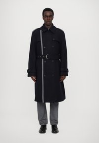 Cappotto trench in lana blu navy con una striscia grigia, design doppiopetto, spalline, dettaglio della cintura e bottoni a contrasto. Indossato con pantaloni grigi.