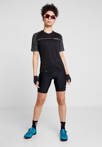Svart sykkeltrøye med korte ermer, aksentuert med oransje sting. Brukt med svarte sykkelshorts og blå sykkelsko, med fingerløse hansker.