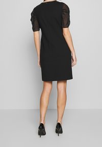 Černé šaty s průhlednými balónovými rukávy, přiléhavý střih a skrytý zip na zadní straně. Nosí se s černými lodičkami na vysokém podpatku.