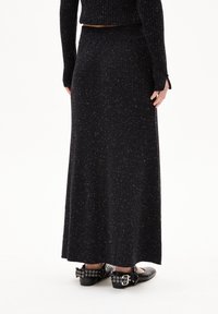 Langer, schwarzer Rippstrickrock mit silbernen Sprenkeln, ausgestattet mit einer taillierten Taille und einem fließenden, knöchellangen Design. Kombiniert mit flachen, schwarzen Schuhen.