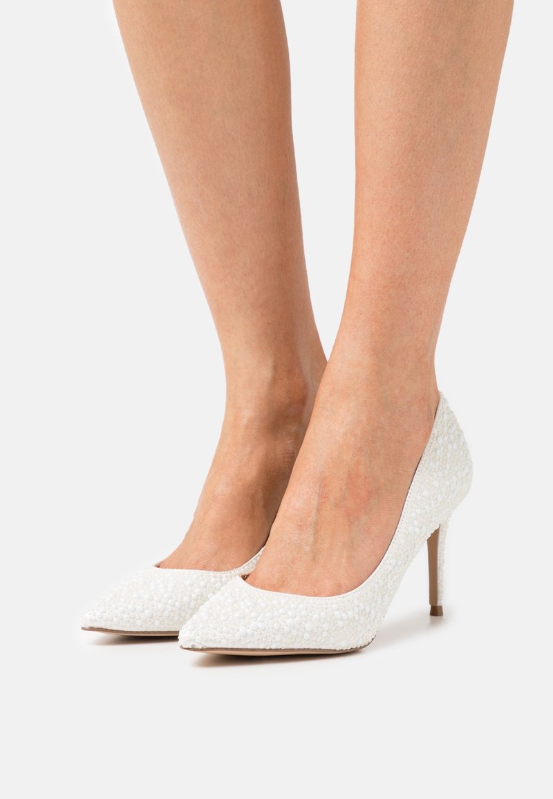 Jambes portant des escarpins blancs ornés de petites perles, bout pointu et talons aiguilles fins sur un fond neutre.