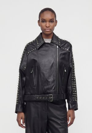 Femme portant une veste de motard en cuir noir avec des clous argentés aux épaules et aux manches, zippée et ceinturée, debout devant un fond uni.