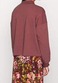 Felpa bordeaux con collo alto, spalle cadenti e polsini a costine, abbinata a una gonna a fiori con tonalità rosse, rosa e verdi.