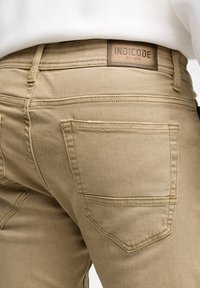 Pantalones beige con una vista en primer plano de la parte trasera, que presenta dos bolsillos traseros y una etiqueta marrón grabada con "INDICODE EST. 1993."