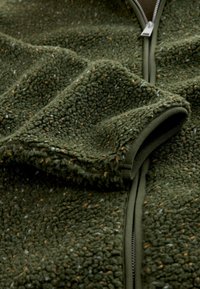 Primo piano del tessuto di una giacca in pile verde con motivo a forma di freccia, trama visibile, polsino e dettaglio della cerniera.