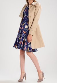 Beige trenchcoat över en blå blommig klänning med volangkant. Klänningen har ett livligt blommönster. Ljusgrå högklackade skor.