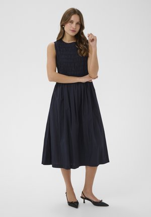 Žena stojaca so skríženými rukami, oblečená v bezrukávových námorníckych midi šatách a čiernych lodičkách so špičatou špičkou na opätku, na bielom pozadí.