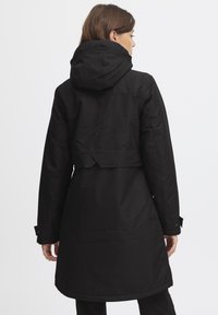 Parka imperméable noire avec capuche ajustée, taille ceinturée et manches longues. Présente une texture lisse et des poches dissimulées.
