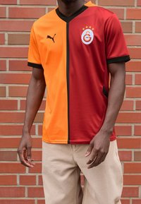 Camiseta de fútbol en naranja y rojo, con una franja vertical negra. Incluye un logo con estrellas y un diseño de cuello en V. Mangas cortas.