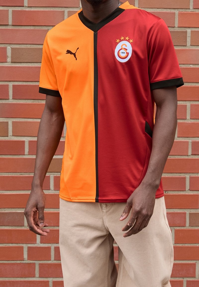 Camiseta de fútbol en naranja y rojo, con una franja vertical negra. Incluye un logo con estrellas y un diseño de cuello en V. Mangas cortas.