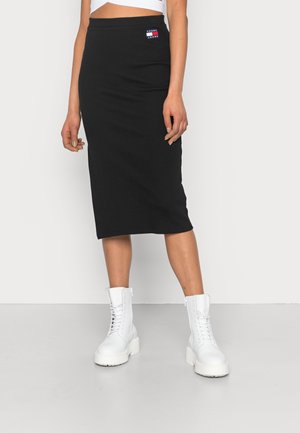 Mujer con una falda midi negra con el logo de Tommy Jeans y botas blancas gruesas con cordones, de pie contra un fondo liso y claro.