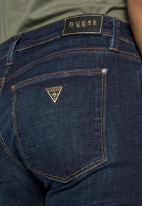 Vaqueros de mezclilla azul oscuro con pespuntes naranjas contrastantes, con un logo triangular en el bolsillo trasero y una etiqueta de marca negra en la cintura.