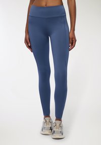 Legginsy w kolorze niebieskim, z wysokim stanem, wykonane z elastycznego materiału, z gładką fakturą i detalami na bocznych szwach. Połączone z beżowymi sneakersami.