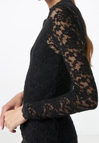 Svart spets långärmad topp med en figurnära design och genomskinliga spetsärmar med blommönster. Slät bodice med rund halsringning.