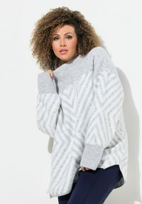 Un pull gris à col montant, orné de motifs en zigzag blancs, avec des poignets côtelés et une coupe ample. Associé à des leggings noirs.