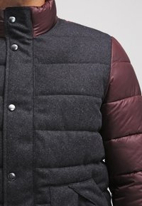 Vadderad jacka med en kropp i en mörkgrå ullblandning och ärmar i glänsande vinröd tyg. Silverfärgade knappringar och en hög krage fulländar designen.