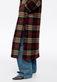 Persona vistiendo un abrigo largo de cuadros rojos, verdes y negros sobre jeans azules holgados y zapatos negros puntiagudos, de pie con las manos en los bolsillos del abrigo.