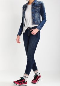 Chaqueta de mezclilla con un lavado azul, camiseta gris claro, jeans ajustados oscuros y zapatillas rosas. Calcetines a rayas visibles por encima de los zapatos.