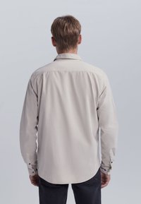 Camisa de manga larga color beige claro con cuello, puños abotonados y dobladillo curvado. Fabricada con una tela suave, exhibe un diseño simple y limpio.