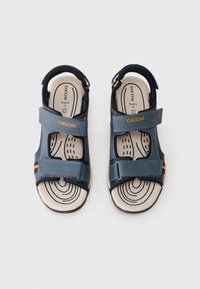 Sinised sandaalid takjakinnitustega, ristikujuline disain, kummist tallad ja beež sisetald lainelise mustriga. Logo asub rihmal.